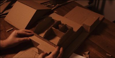 Film still from '3 Barley Road' (2024) by Jon Beaton. A scale model of a bungalow is being constructed out of cardboard. Two hands are placing a piece of the model.
